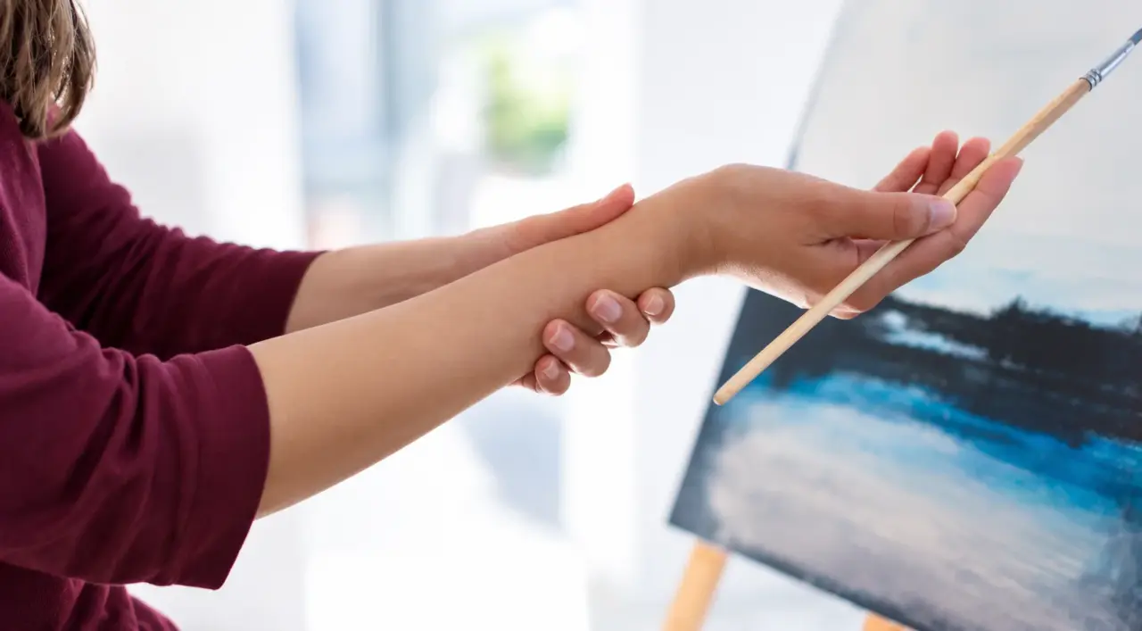 Therapist supporting a patient&rsquo;s wrist while she holds a paintbrush at an easel