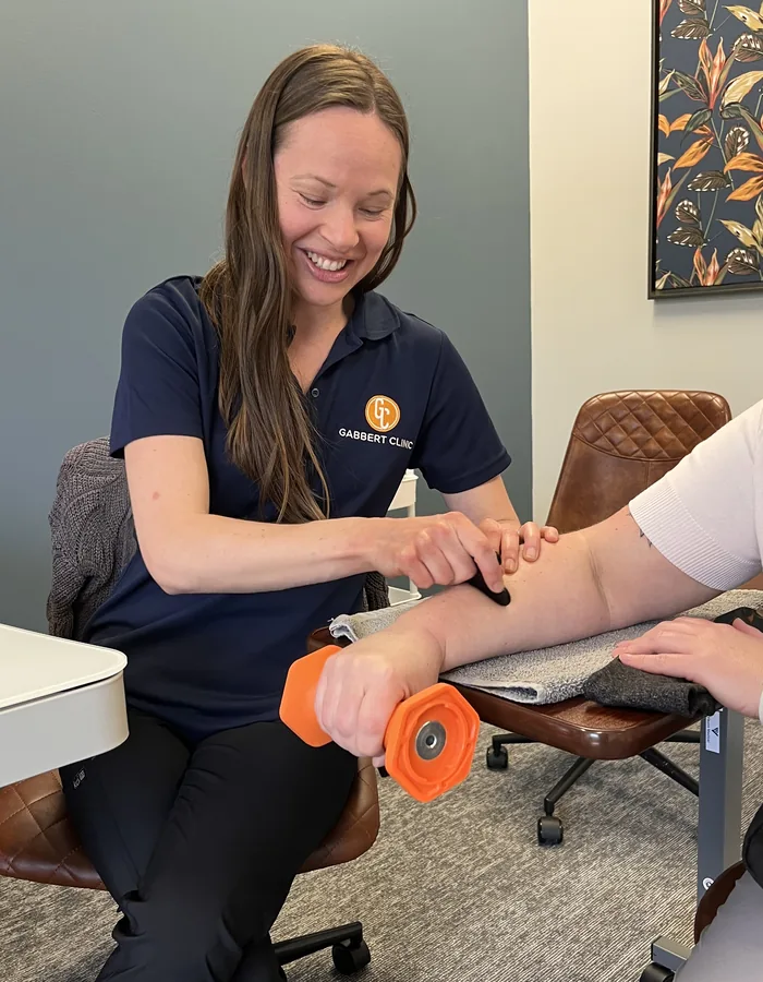 A smiling Gabbert Clinic therapist uses a soft tissue tool on a patient's forearm while the patient holds a light hand weight