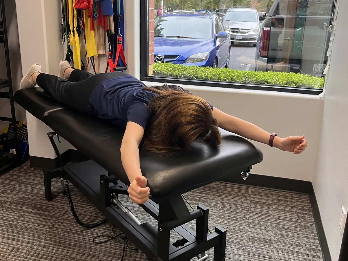 Patient lying face down on a treatment table with both arms extended out to the sides during a shoulder exercise