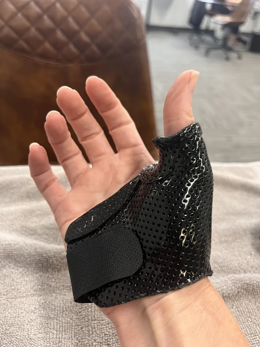Black perforated thermoplastic thumb splint molded around the base of the thumb on an open palm, held with a strap