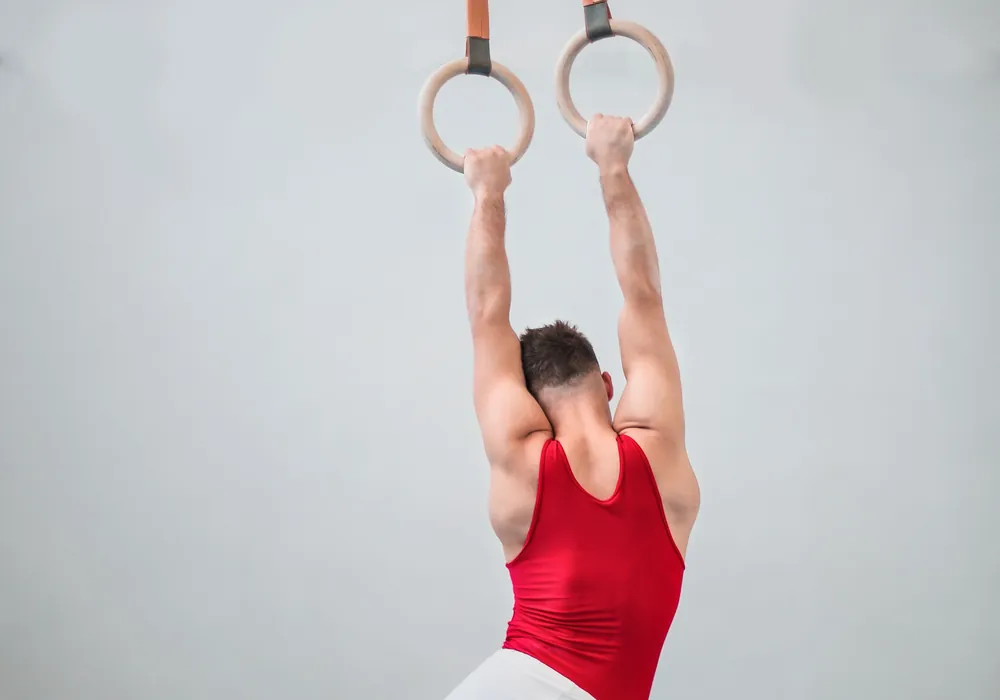 Gymnast in a red leotard hanging from wooden still rings with both arms extended overhead